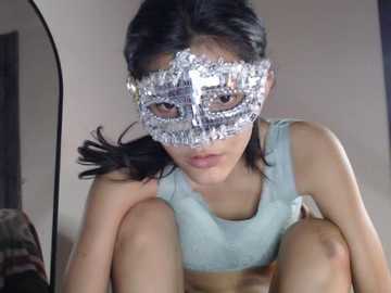 Video of an Asian woman with long black hair, wearing a silver masquerade mask and light blue tank top, sitting in a dimly lit room, with a mirror reflecting part of her body.