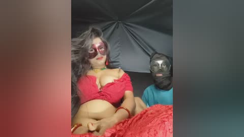 Video of a woman in a red, off-the-shoulder top with large breasts and a man in a black mask, sitting on a red bed.