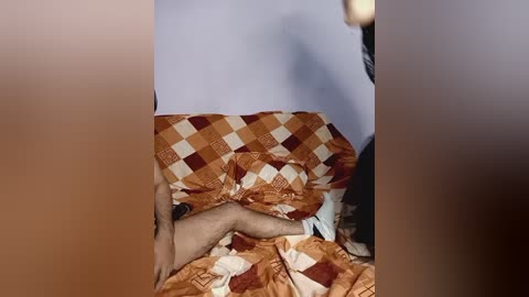 Video of a man lying on a bed with a patterned orange and brown quilt, partially covered by another person, who is partially visible in the background.