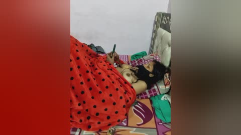 Video of a dark-haired woman with medium breasts, lying on a bed under a red polka-dot blanket, holding a smartphone, surrounded by colorful bedding and a white wall.