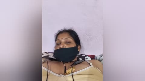 Video of an Asian woman with medium skin tone, tied to a white wall, wearing a yellow tank top and black face mask, with visible distress and fear.