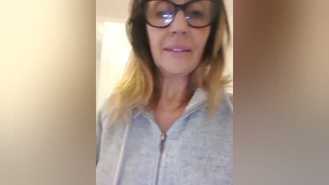 Media: Video of a middle-aged woman with fair skin, shoulder-length blonde hair, and large black-framed glasses, wearing a light grey zip-up hoodie, smiling slightly. Background is blurred and beige.