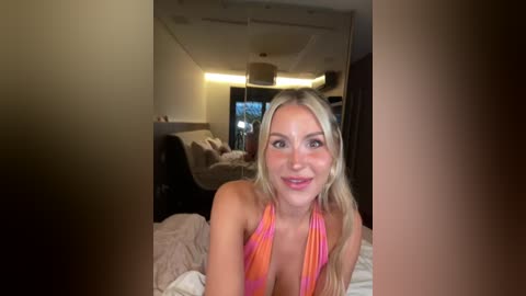 Video of a blonde woman in a colorful halter top, smiling in a dimly lit, modern bedroom with a large TV and bed.