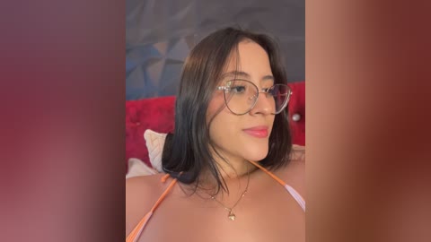 Video of a young woman with medium skin tone, straight black hair, and glasses, wearing an orange halter top, sitting on a red bed with a geometric-patterned wall in the background.