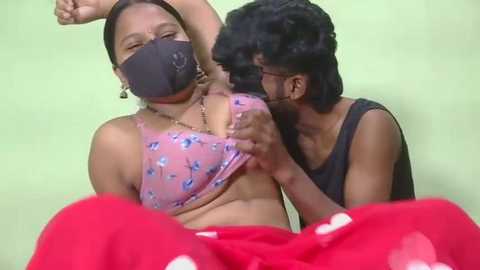Video of a young woman in a pink bra with blue flowers, red pants, and a black face mask, being kissed passionately by a man in a black tank top and glasses.