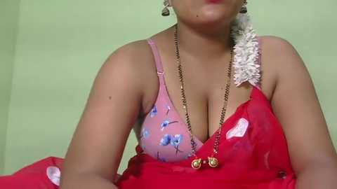 Video of a woman with light brown skin, wearing a pink floral bra and a red saree with a white floral hair accessory, sitting against a green wall.
