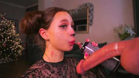 Video of a young woman with fair skin, brown hair in a bun, wearing a black lace top, kissing a large, erect penis. Background includes a Christmas tree and framed mirror.