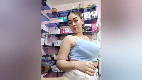 Video of a young woman with light brown skin, medium build, and straight black hair in a ponytail, wearing a light blue tank top and white shorts, standing in a cluttered, brightly lit pharmacy.