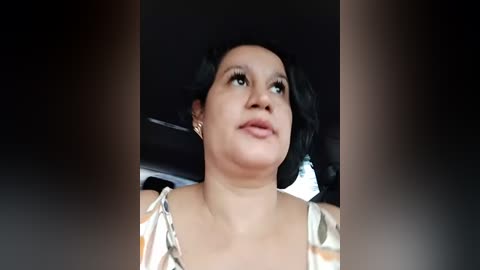 A video of a woman with short, wavy black hair, wearing a white and black polka-dotted blouse, sitting in a car. The background is dark, and she appears thoughtful.