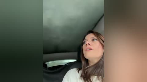 A video of a woman with fair skin and long brown hair, wearing a white top, gazing upwards with a thoughtful expression, inside a car with a muted, green-tinted interior.