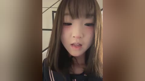 Video of a young Asian girl with light skin, straight brown hair, and bangs, blushing slightly, wearing a dark blue shirt. Background is a blurred indoor setting with beige walls.