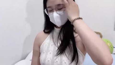 Media: Video of an Asian woman with long black hair, wearing a white mask, glasses, and a white knit top, touching her forehead. Background shows a white wall and a green stuffed toy.