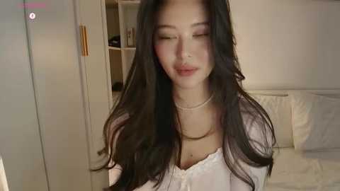 Media: Video of an Asian woman with long, black hair, wearing a white blouse and pearl necklace, smiling gently in a softly lit bedroom.