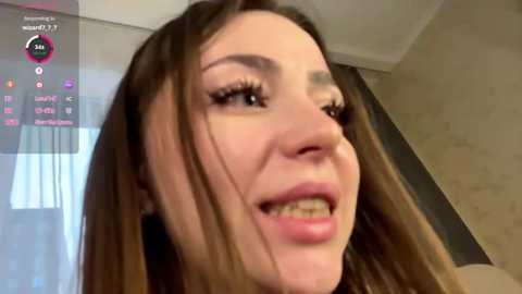 Video of a smiling, young woman with long, brown hair, wearing makeup, including long eyelashes, in a modern indoor setting with a large window and tiled wall.