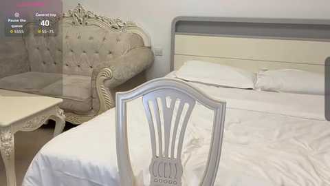 Video of a modern, minimalist bedroom with a white bed, cream upholstered bench, and a vintage-style wooden chair. The bed has a simple headboard, and the room features soft, neutral tones and clean lines.