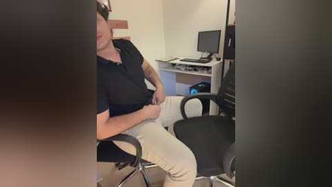 Video of a man in a black polo shirt and beige pants sitting in a black office chair, facing a white desk with a monitor and keyboard, in a minimalist office setting.