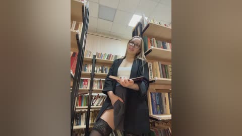 Video of a blonde woman in glasses, wearing a black blazer over a white shirt, black skirt, and thigh-high stockings, standing in a book-filled library, holding a book and a pen, looking thoughtful.