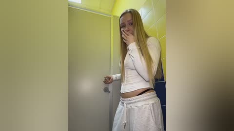 Video of a young blonde woman with long hair, wearing a white long-sleeve crop top and matching pants, standing in a yellow-tiled bathroom, looking shocked and holding her mouth.