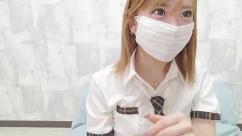 Video of a young Caucasian woman with light brown hair, wearing a white button-up shirt, a striped tie, and a white surgical mask, seated in a room with white patterned wallpaper.