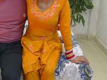 Video of a woman wearing an orange kurta with pink embroidery, seated next to a man in a pink shirt on a white and blue patterned couch.