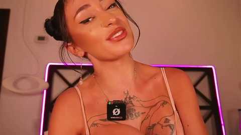 Video of a smiling, dark-haired woman with a tan, wearing a camo top and a dog tag necklace, showcasing a tattoo on her chest, against a pink-lit background.