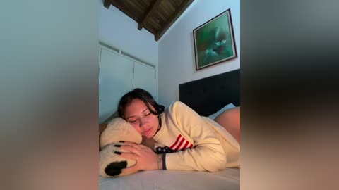 Video of a young woman lying on a bed, wearing a striped shirt, holding a teddy bear, in a cozy bedroom with wooden ceiling beams and framed art.