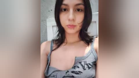 Video of a young, light-skinned woman with shoulder-length black hair, wearing a grey tank top with a graphic design, standing indoors against a textured white wall. She has a small facial mole on her right cheek.