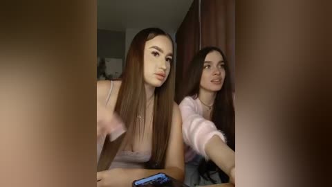 Video of two young women, one with long brown hair in a pink top, the other with long black hair in a purple top, leaning over a table, blurry background.