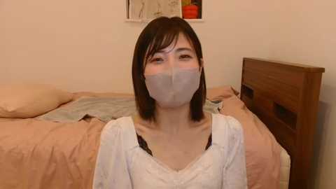 Media: Video of an East Asian woman with short black hair, wearing a white long-sleeve blouse, black bra, and a gray face mask, sitting on a beige bed with a wooden headboard in a simple, beige-walled room.