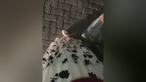 A video captures a person from behind, wearing a white dress with black floral patterns, standing on a textured stone pavement. The background is dimly lit, creating a moody, slightly blurred atmosphere.