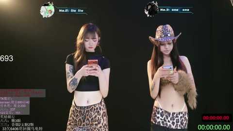 Video of two young women with fair skin, one with a black crop top and leopard print skirt, the other in a leopard print hat and shorts, both holding phones.