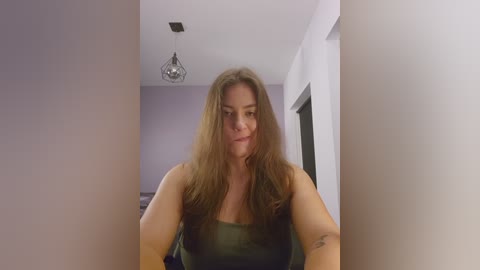 Video of a young woman with long, wavy brown hair, wearing a dark green strapless top, standing indoors. Background features a white ceiling with a hanging light fixture and white walls.