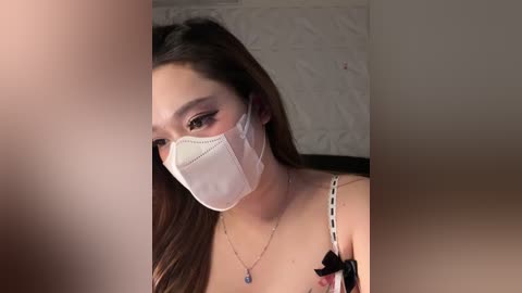 Video of a young woman with long dark hair, wearing a white face mask and black lace bra. She has fair skin, brown eyes, and a delicate necklace. Background shows a textured wall.