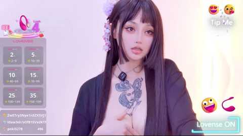 Video of a young East Asian woman with long black hair, pink floral hairpiece, and a black open jacket revealing a large tattoo on her chest. She's in a livestream setting with a colorful calendar overlay.