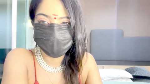 Media: A close-up video of a woman with long black hair, wearing a black face mask, red bindi, and a silver necklace, sitting in a modern, minimalistic room with a gray couch.
