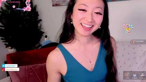Video of a smiling Asian woman with long black hair, wearing a teal sleeveless dress, sitting on a red couch, surrounded by a virtual background with colorful hearts.