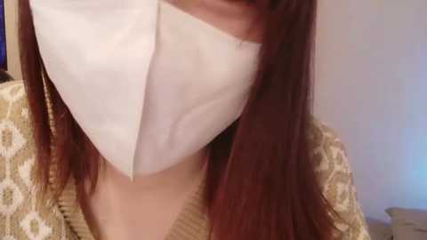A video shows a person with long, straight, dark red hair wearing a white face mask and a beige, patterned cardigan. The background is out of focus, suggesting an indoor setting.