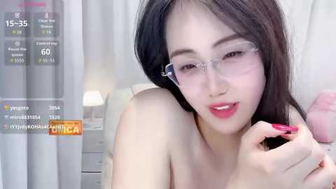 Video of a young East Asian woman with fair skin, long black hair, and clear glasses, applying red lipstick in a soft-focus indoor setting.