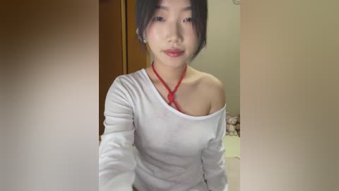 Media: Video of a young Asian woman with fair skin and black hair in a white off-shoulder top and red choker. She has a neutral expression. Background shows a beige wall and a wooden door.