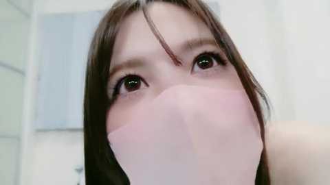 Video of a young Asian woman with straight brown hair, large brown eyes, and a pink face mask covering her nose and mouth, taken from a low angle, with a blurred, pale background.