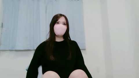 Media: Video of a young Asian woman with long black hair, wearing a black long-sleeve top and pink face mask, sitting on a bed in a white room with light blue curtains.