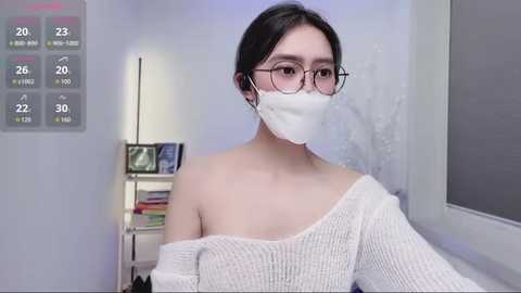 Video of an East Asian woman with glasses and a white mask, wearing a white off-shoulder sweater, sitting indoors with a thermometer showing 28\u00b0C.