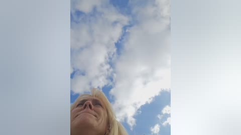 Media: Video of a blonde woman with a slight smile, looking upwards at a bright blue sky with fluffy white clouds.