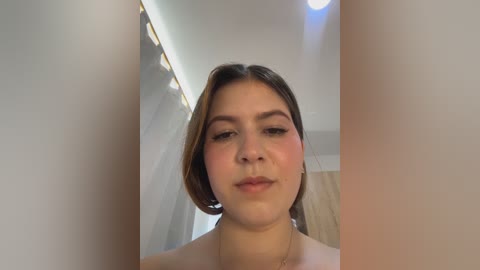 Video of a young woman with light brown skin, short dark hair, and rosy cheeks, standing in a modern bathroom with a white ceiling and wooden cabinet.