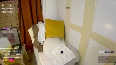 A video of a cozy, neatly made bed in a corner, adorned with a mustard-yellow pillow, a beige blanket, and a white sheet, set against white walls and wooden accents.