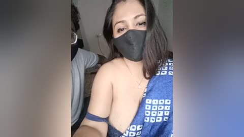 Video of a young Asian woman with medium skin tone, black hair, wearing a black mask, blue patterned dress, and a blue armband, taken indoors, blurred background.