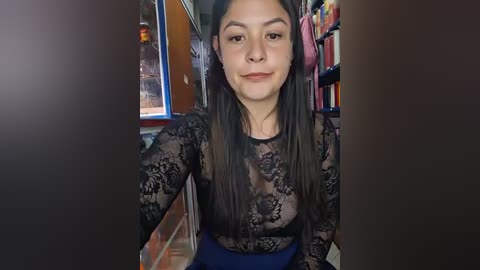A video of a young Latina woman with long dark hair, wearing a black lace top and blue skirt, sitting in a cluttered room with bookshelves and a glass cabinet.