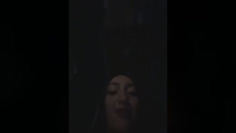 A dimly-lit video of a woman's face, partially obscured by a black vertical frame, with a dark, shadowy background. Her eyes are closed, and her expression is serene.