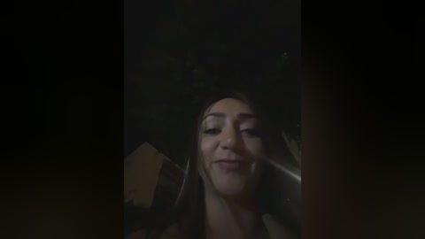 A dimly-lit, low-resolution video shows a woman with long brown hair, wearing a jacket, smiling slightly. The background is dark and blurry, suggesting an outdoor night scene.
