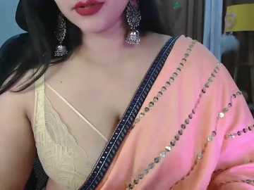 Media: Video of a South Asian woman in a beige blouse with lace detailing, wearing a pink saree with silver embroidery and large, ornate earrings, smiling with red lipstick.